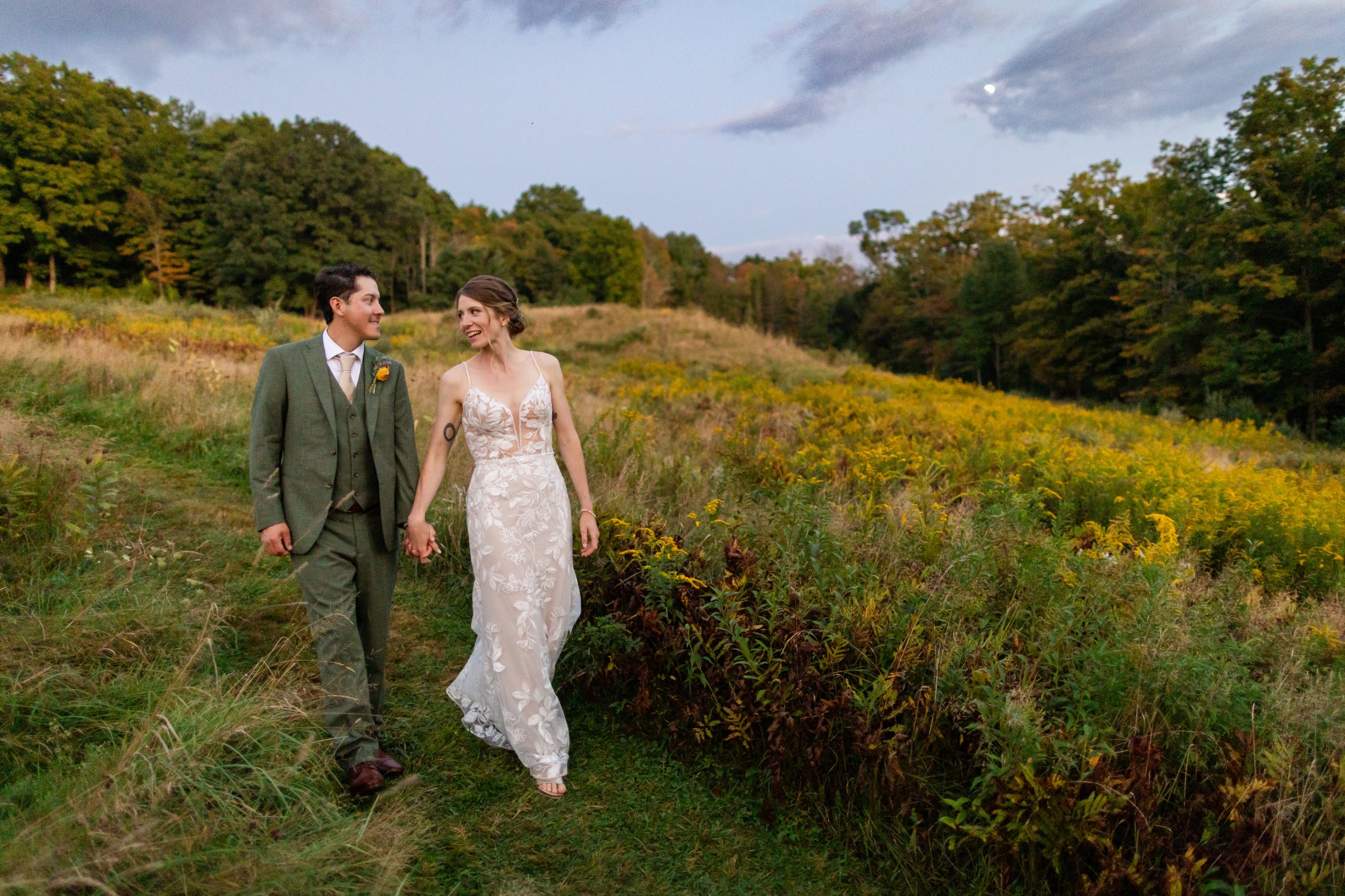 A couple in formal attire walks hand in hand through a grassy field beneath a cloudy sky, at the Montague retreat center in Montague, Massachusetts.