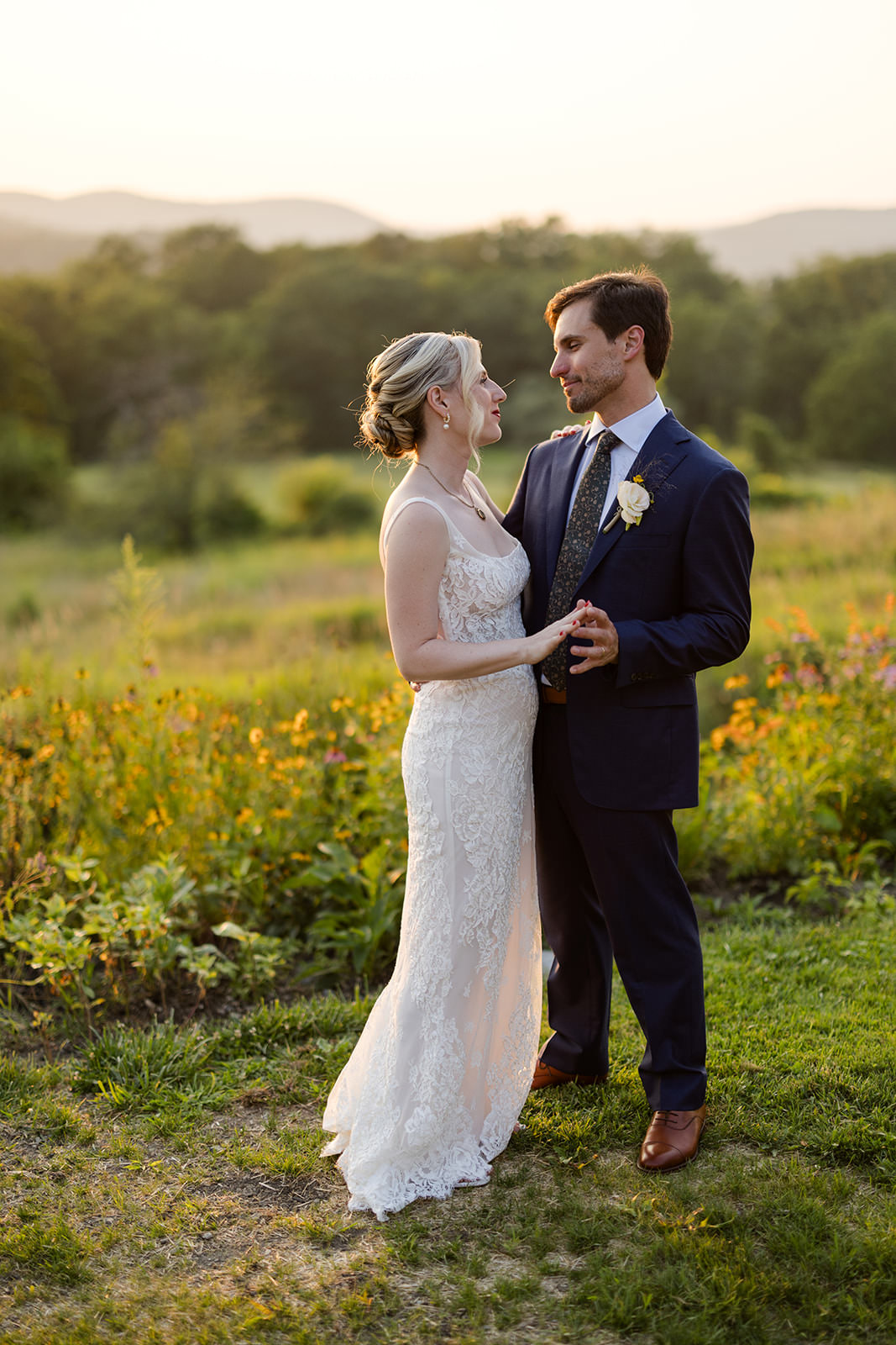 A bride and groom stand outdoors in formal attire, holding hands and looking at each other, with a field and hills in the background—captured beautifully by a Western Mass wedding photographer.