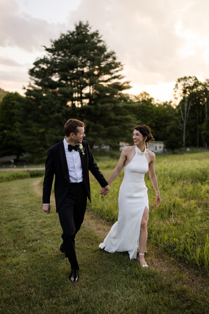 A couple in formal wedding attire holds hands and walks through a grassy field at sunset, smiling at each other, beautifully captured by a Western Mass wedding photographer.