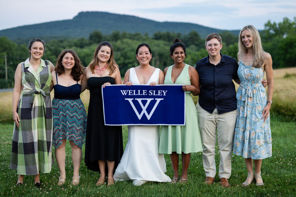 Seven people standing on grass, smiling, with one in a white dress holding a blue “WELLE SLEY W” banner; trees and a mountain are visible in the background.