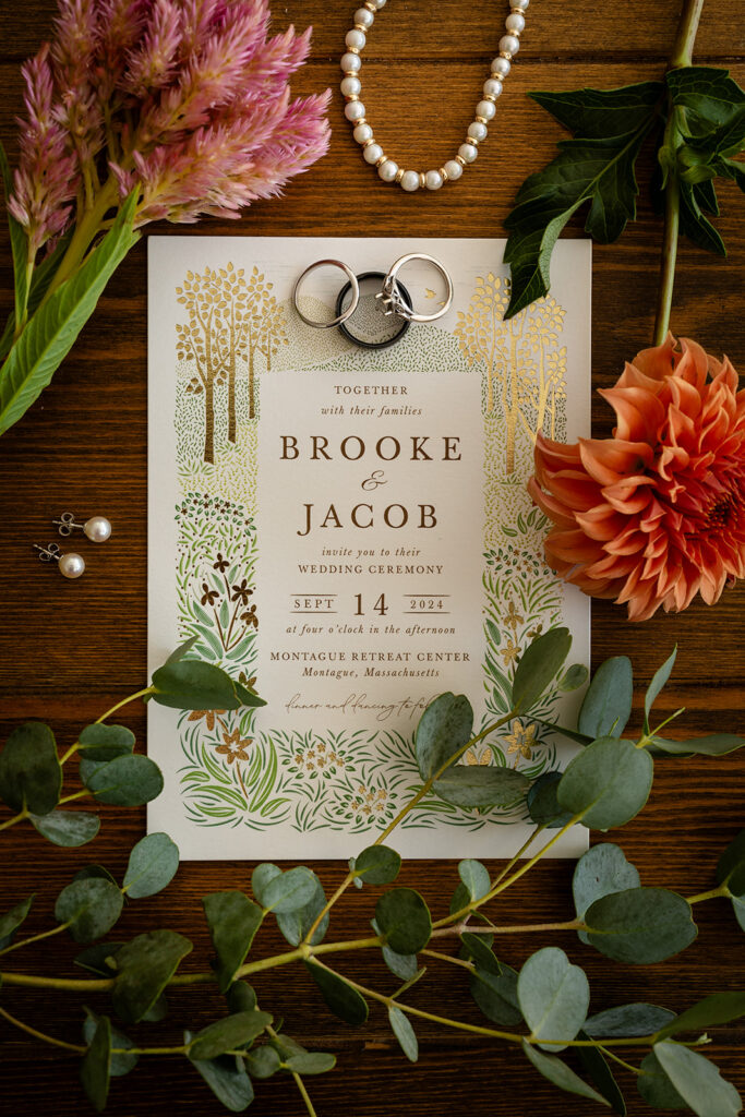 A wedding invitation with floral and forest designs is surrounded by flowers, foliage, pearl jewelry, earrings, and three rings on a wooden surface.