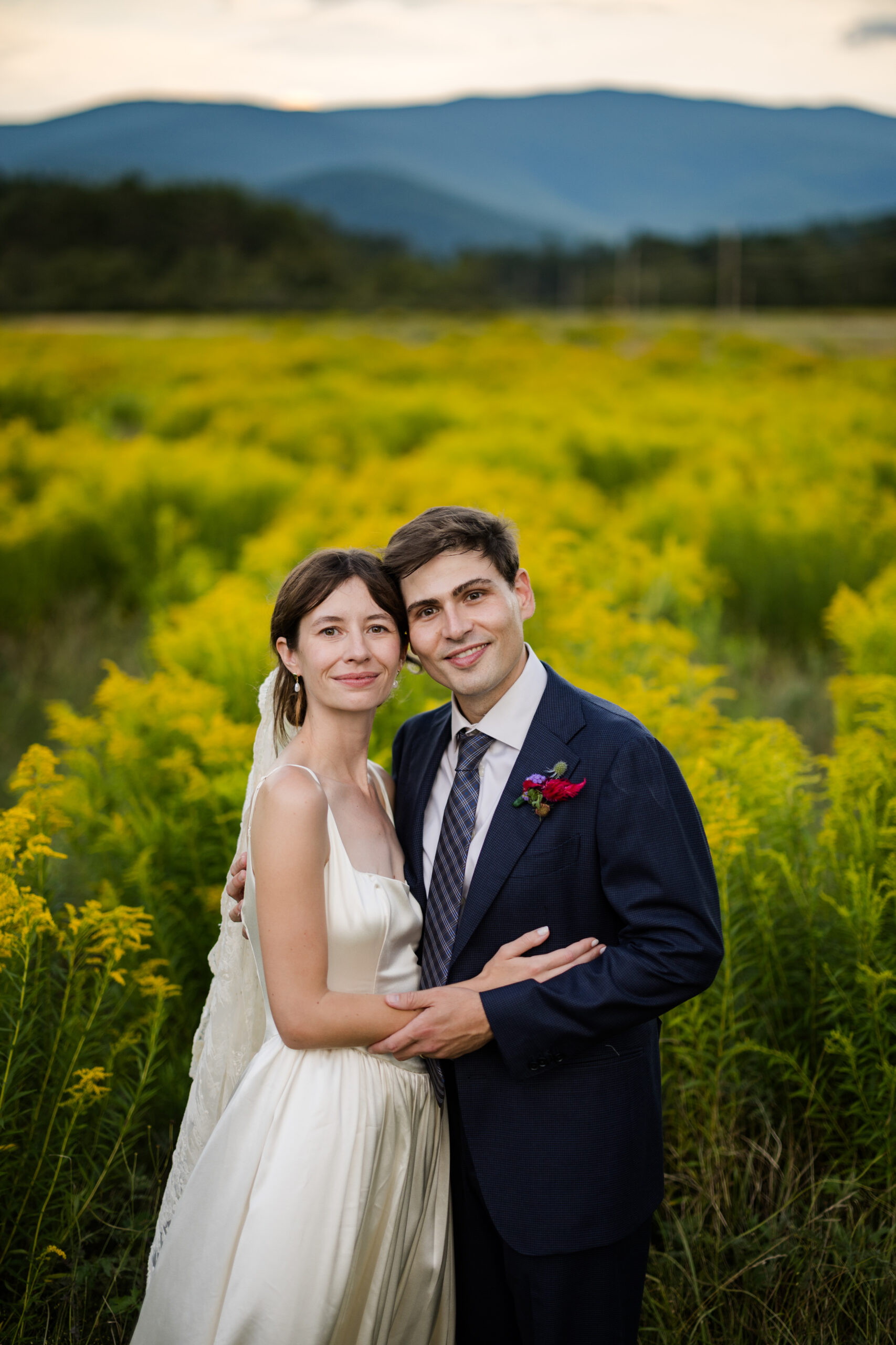 Bride in a white dress and groom in a suit pose together in a field of yellow flowers with mountains in the background.