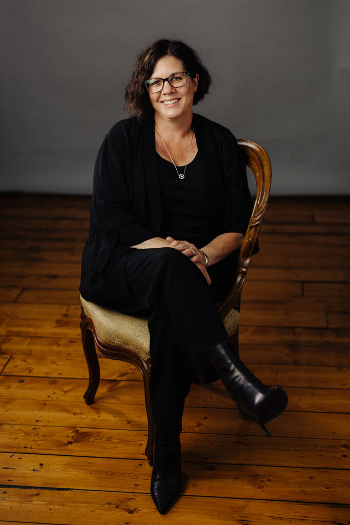 A woman with short dark hair, glasses, and black clothing sits on a wooden chair with legs crossed, smiling, on a wooden floor with a neutral gray background.