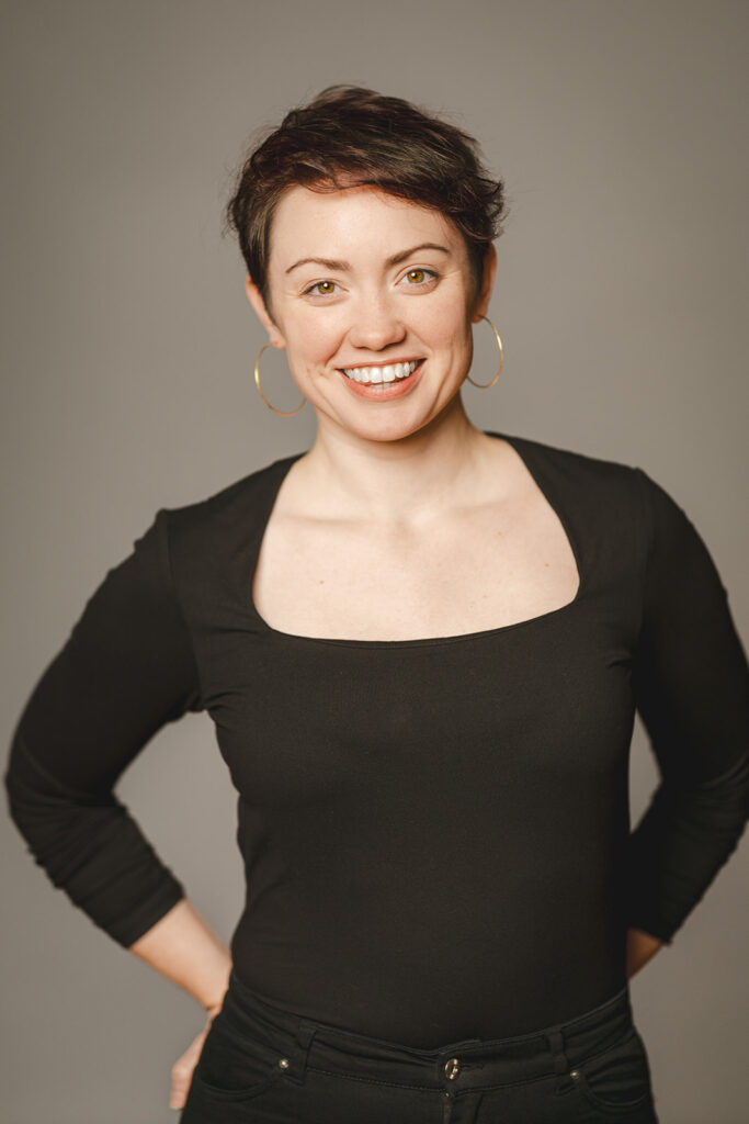 A person with short dark hair, large hoop earrings, and a black square-neck top smiles at the camera against a plain gray background.