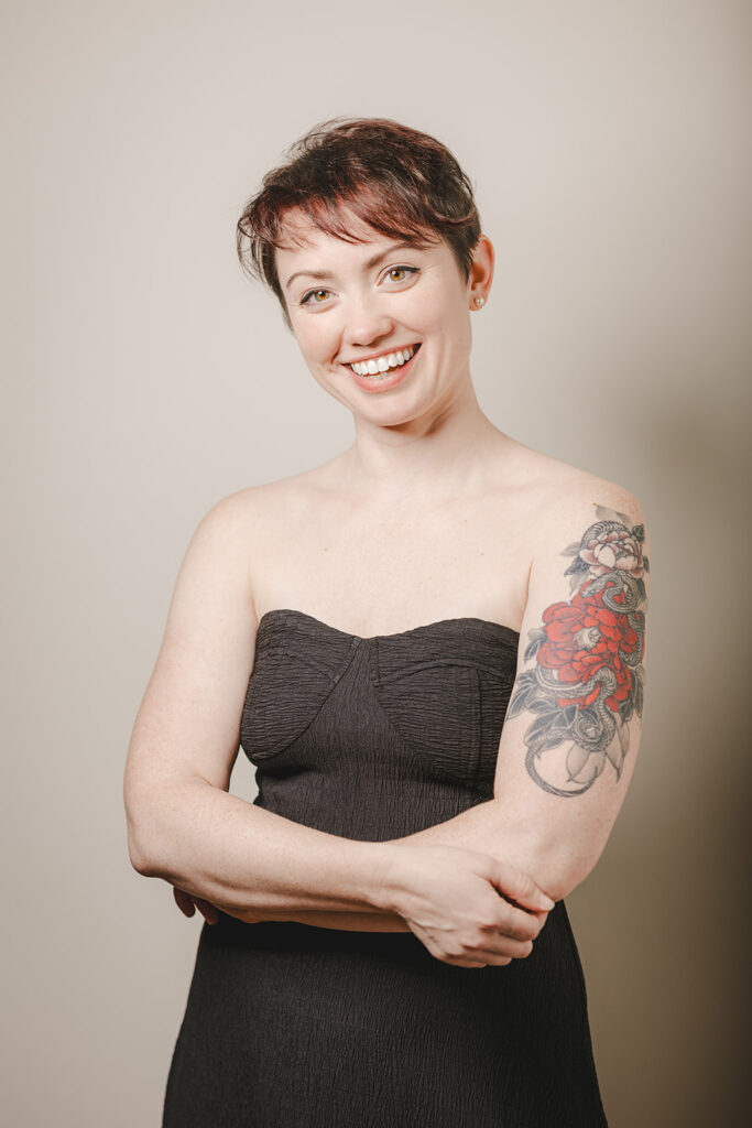 Woman with short hair and a floral tattoo on her upper arm, wearing a strapless black dress, smiles with her arms crossed against a plain background.