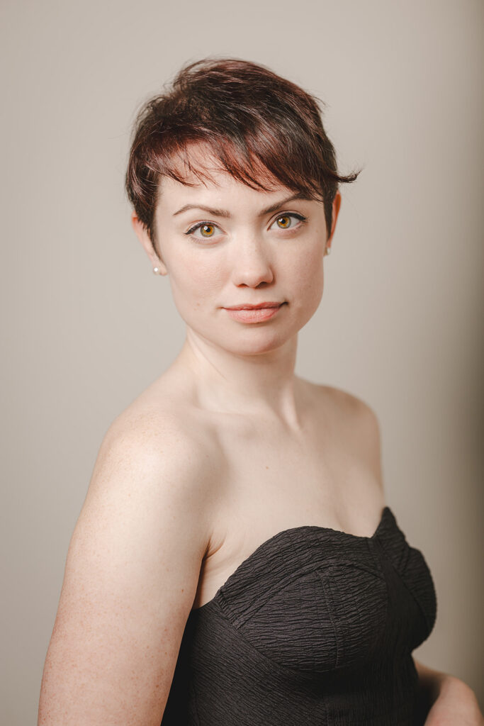 A woman with short brown hair and hazel eyes wears a black strapless top, standing against a plain beige background.