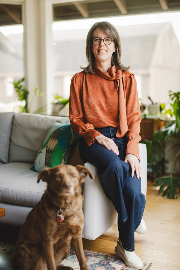 A woman with glasses sits on a couch in a living room, smiling at the camera, with a brown dog sitting on the floor in front of her.