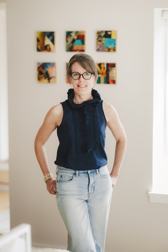 Woman with glasses and a navy sleeveless blouse stands indoors, smiling, with hands in light-wash jean pockets; six small colorful art pieces hang on the wall behind her.