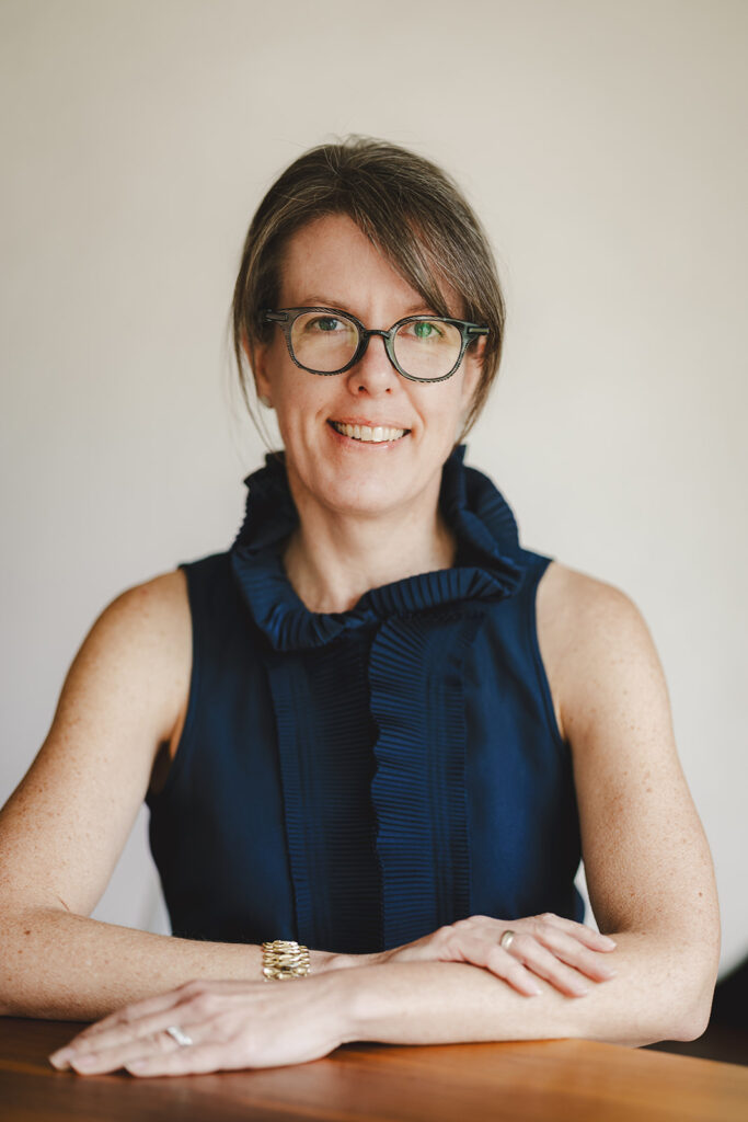 A woman with glasses and a navy sleeveless top sits at a wooden table, smiling at the camera with her arms folded.