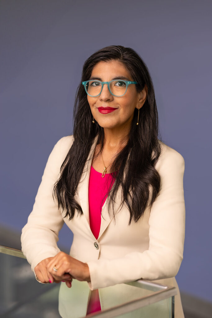 A woman with long dark hair and blue glasses, dressed in a cream blazer and red top, stands indoors in front of a blue background, resting her hands on a glass surface.