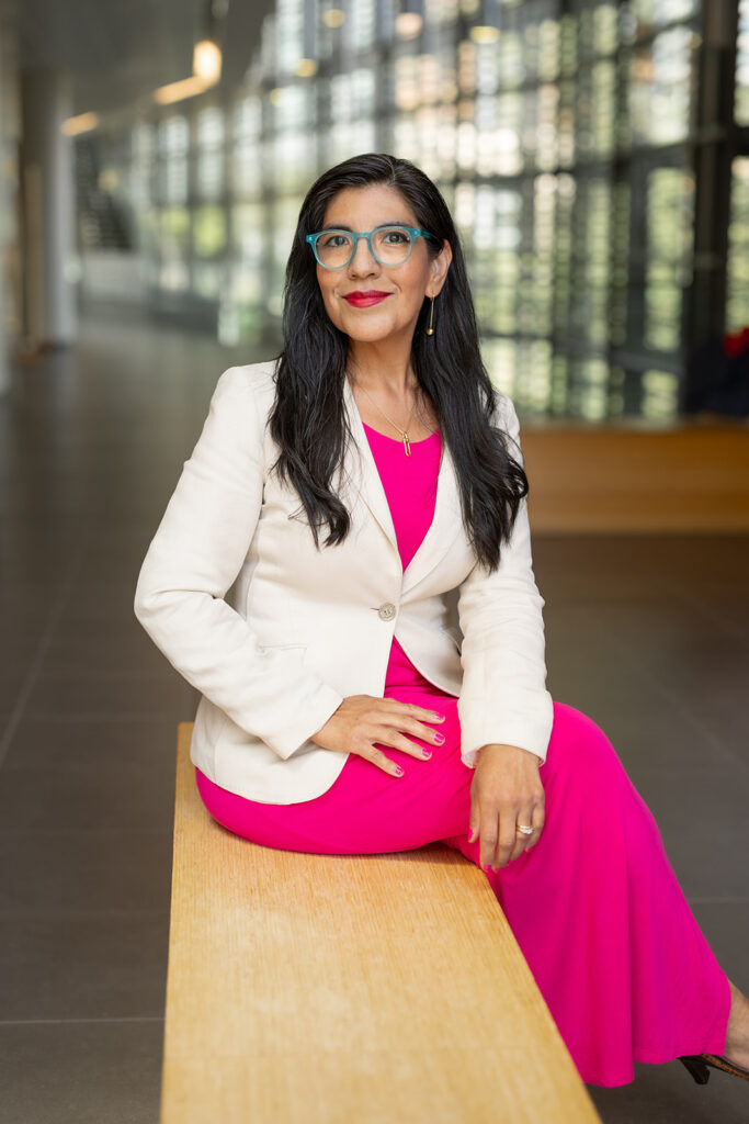 Woman with long dark hair, wearing a white blazer and bright pink dress, sits on a wooden bench in a modern building with large windows.