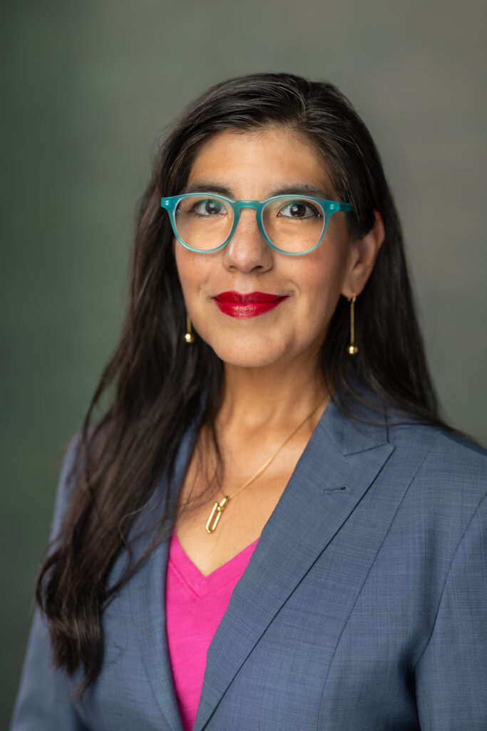 Woman with long dark hair, teal glasses, red lipstick, and a blue blazer over a pink top, poses against a neutral background.