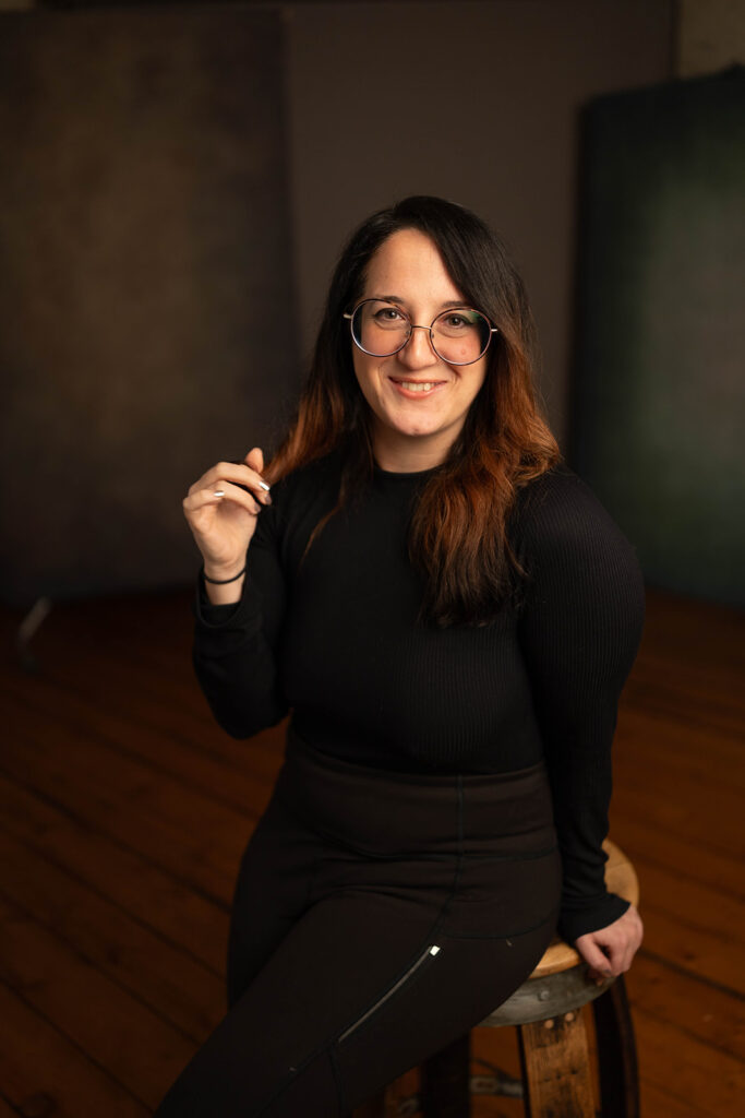 Woman with long dark hair and glasses, wearing a black outfit, sits on a wooden stool in a studio with a brown and green background.