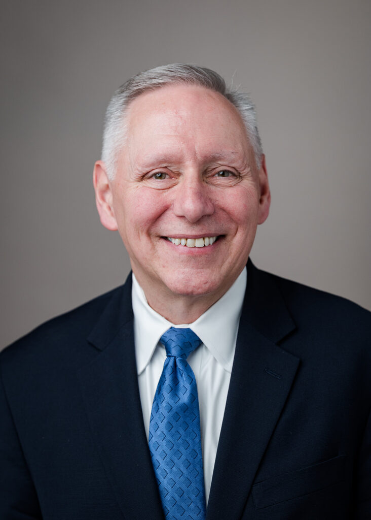 Smiling older man with short gray hair, wearing a dark suit, white shirt, and blue patterned tie, poses for a professional headshot against a plain gray background.