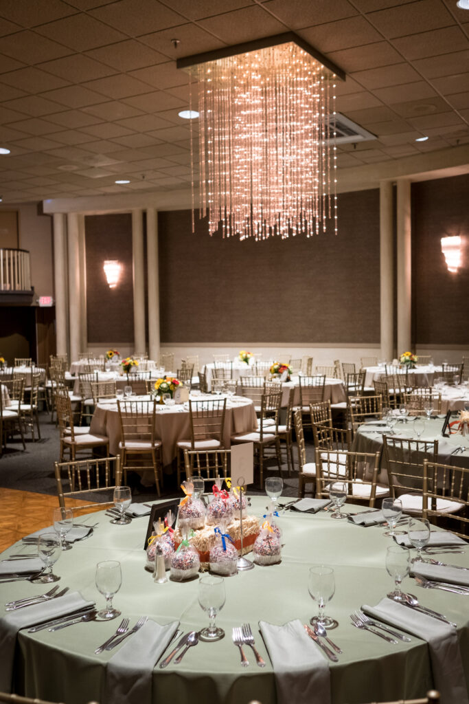Banquet hall with round tables covered in sage green cloths, decorated with flowers and candy centerpieces, under a hanging chandelier.