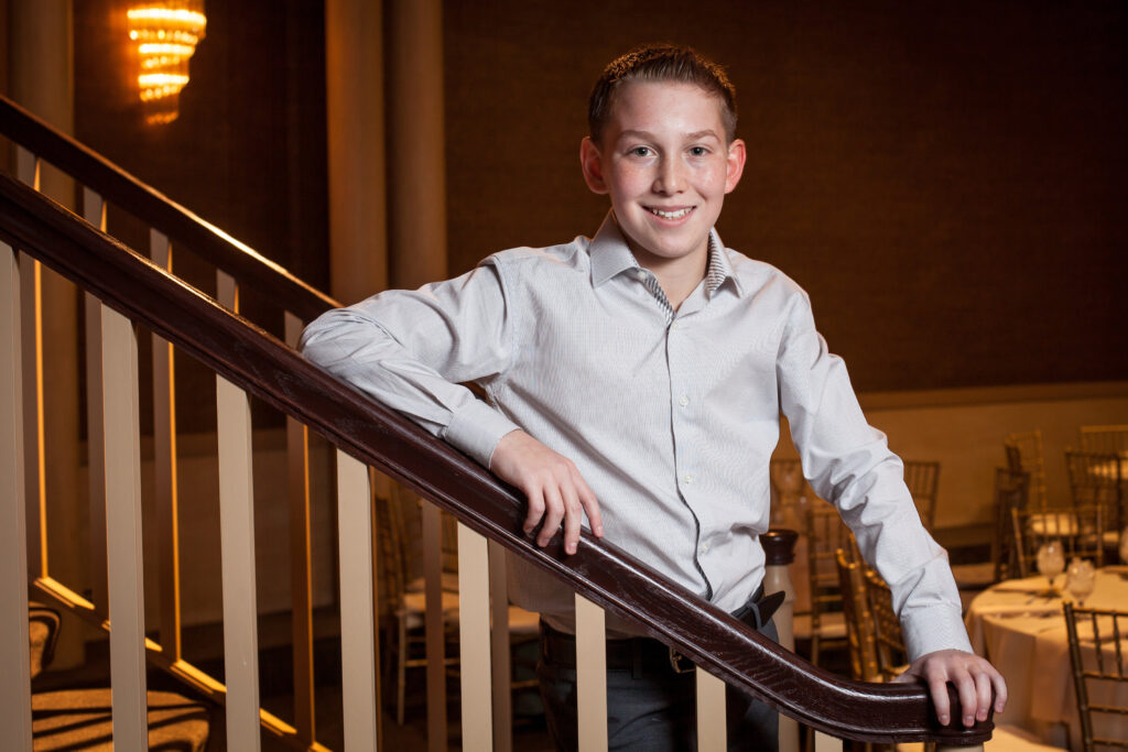 Teenage boy in a light dress shirt stands smiling at the bottom of a staircase in a formal dining room with round tables and gold chairs.