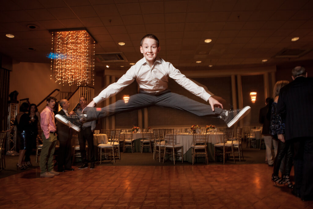 A boy in a white shirt and gray pants performs a mid-air split jump on a dance floor at an indoor event, with people and tables in the background.
