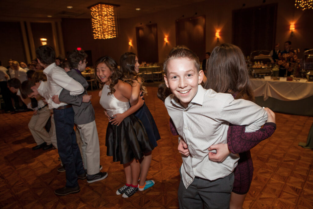 A group of teens stand back-to-back in pairs at a formal event, smiling and posing for a photo on a patterned dance floor with a buffet table in the background.