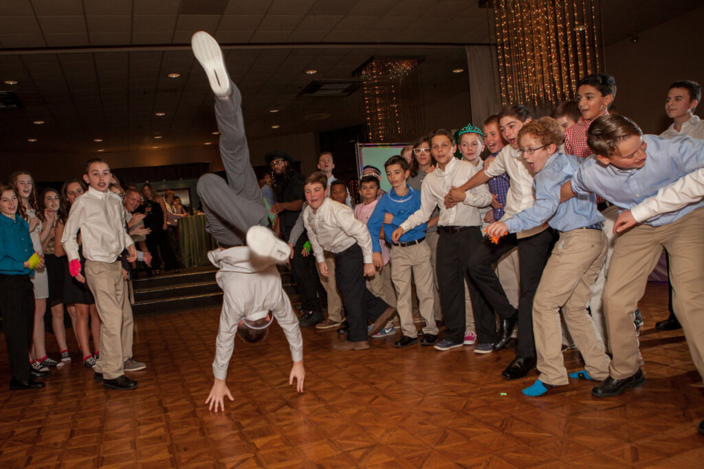 A group of children in dress clothes watch as one boy does a cartwheel on a wooden dance floor at an indoor event.