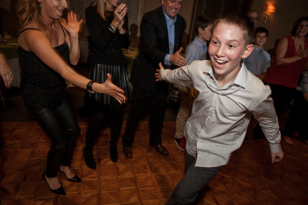 A boy in a light shirt runs and smiles on a dance floor while people around him clap and cheer.