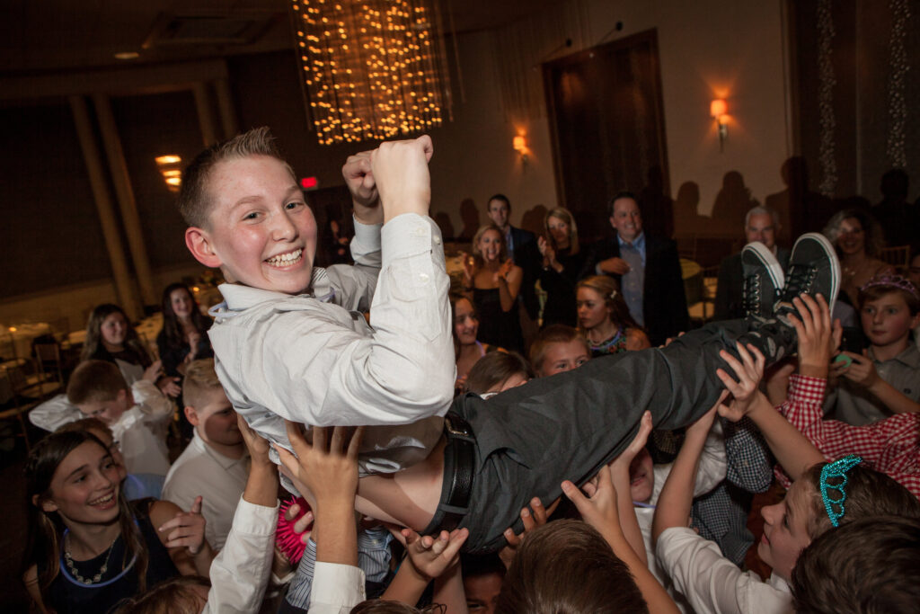 A smiling boy in a dress shirt is being lifted by a cheering group at an indoor party with string lights and guests in the background.