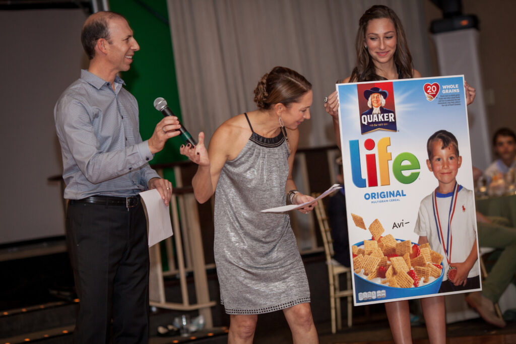 Three adults stand indoors, one holding a microphone, while another holds a large mock-up box of Life cereal featuring a photo of a young boy labeled “Avi.”.
