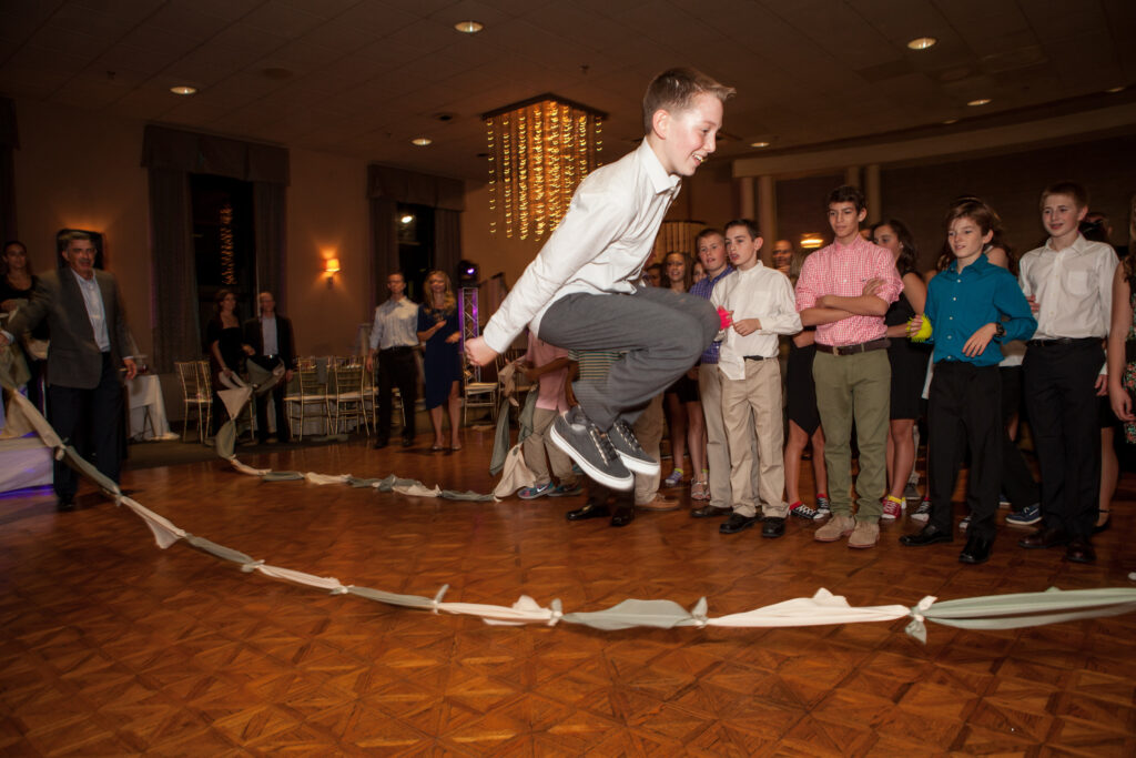 A boy jumps over a long limbo stick in a decorated ballroom as a group of children and adults watch.
