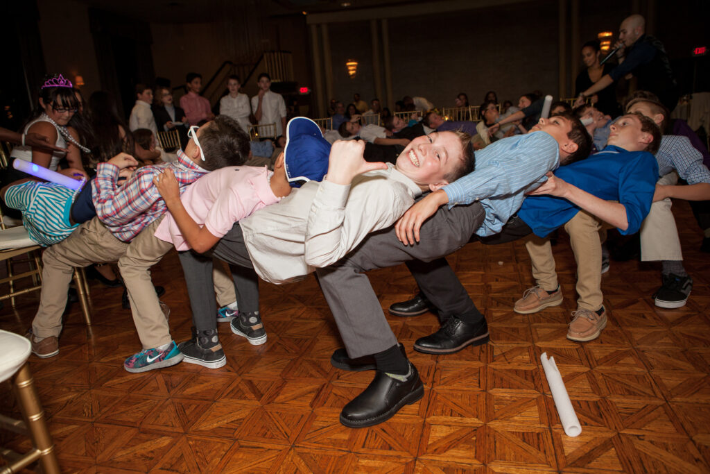 A group of boys at a party are sitting in each other’s laps in a circle, forming a human chair and laughing, on a wooden dance floor.