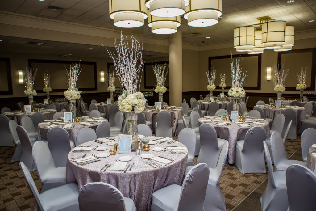 Banquet hall with round tables covered in silver cloth, white floral centerpieces, and gray chairs arranged for an event.