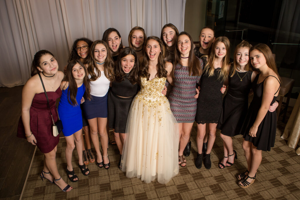 A group of teenage girls pose together indoors, with one girl in a formal cream dress standing in the center, smiling at the camera.