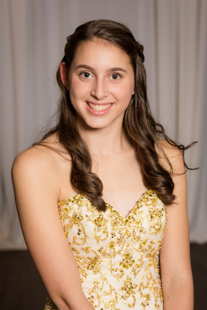 Young woman in a gold-embroidered strapless dress smiles at the camera, posed in front of a plain, light-colored curtain backdrop.