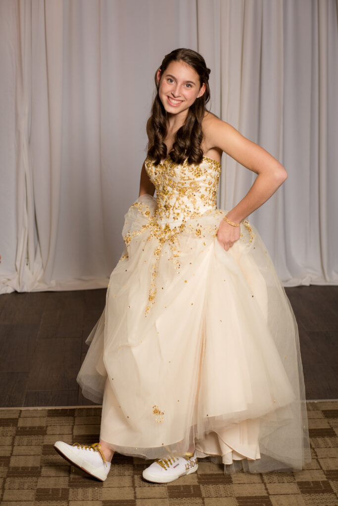 Young woman in a gold-embellished formal gown stands indoors, smiling at the camera, and wearing white athletic shoes with gold laces.