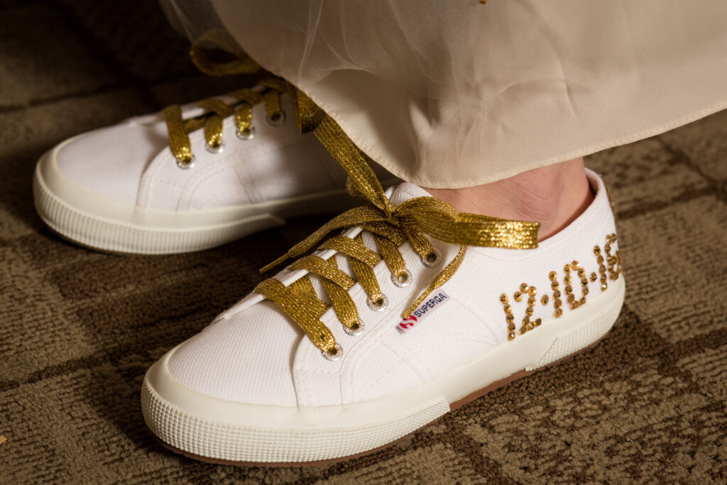 Person wearing white canvas sneakers with gold glitter laces. One shoe has the date “12.10.23” made with gold beads. The person is standing on a carpet, partially covered by a sheer white fabric.