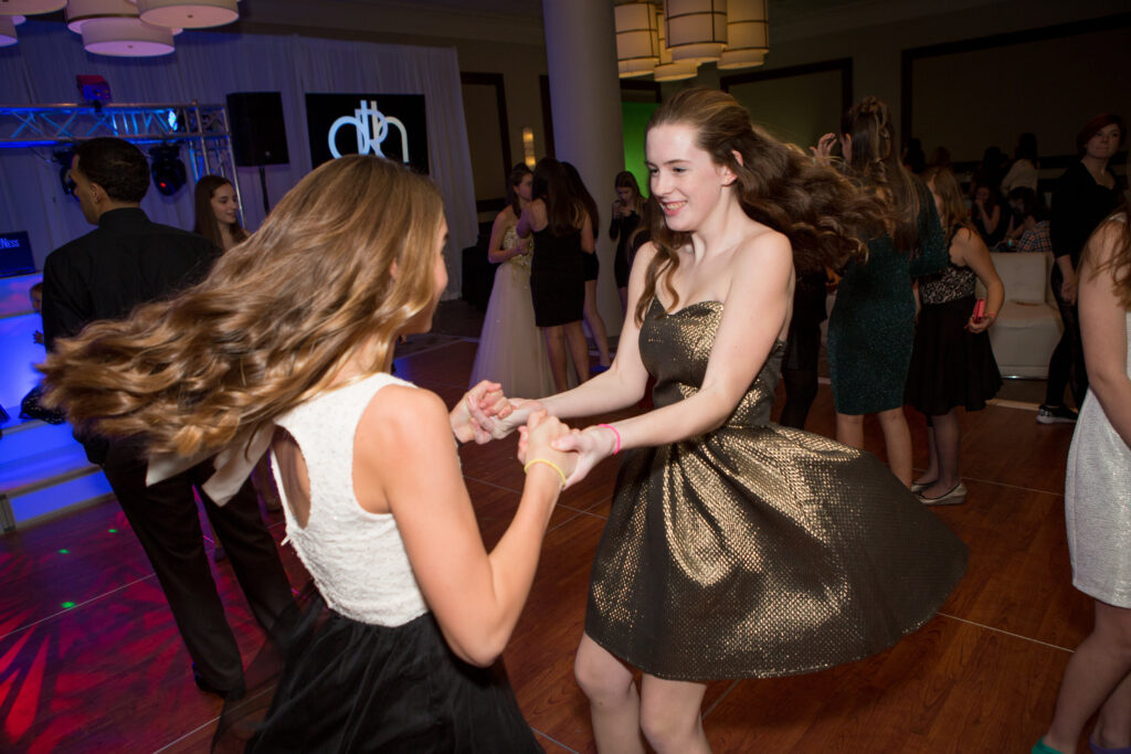 Two young women in dresses hold hands and dance on a wooden floor at a lively indoor event with other people and a DJ in the background.