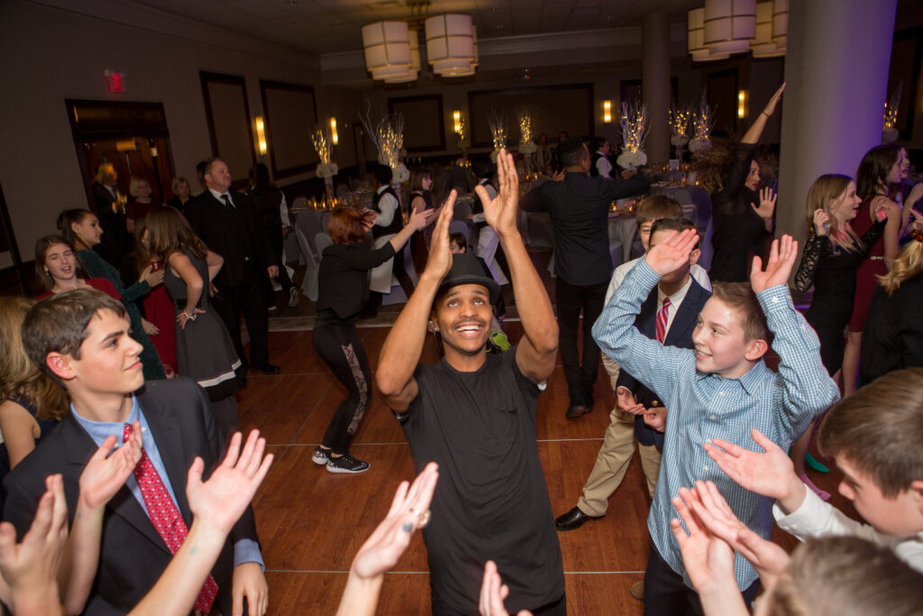 A group of people, including children and adults in formal attire, dance and raise their hands together on a wooden floor at an indoor event.