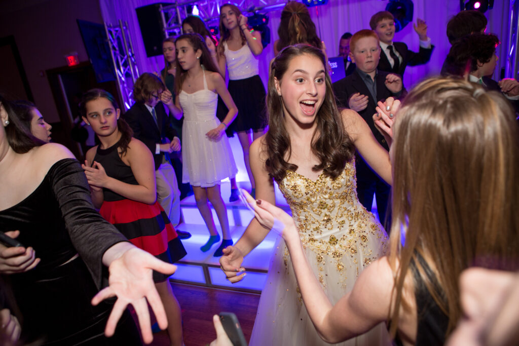 Teens in formal attire dance and socialize at a party with purple lighting and a lively atmosphere.