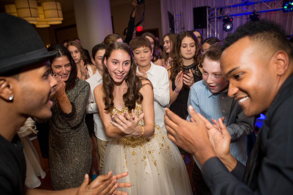 A group of teenagers and adults in formal attire are clapping and smiling together at an indoor party or celebration.