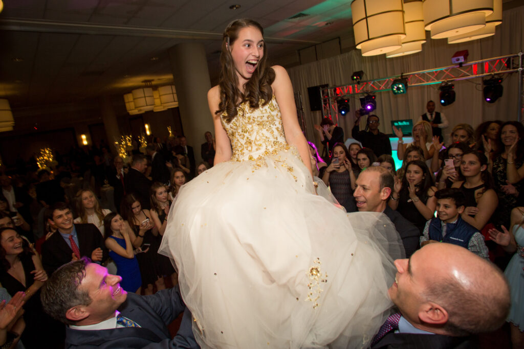 A young woman in a white and gold dress is lifted on a chair by two men at a crowded indoor celebration, with guests watching and taking photos.