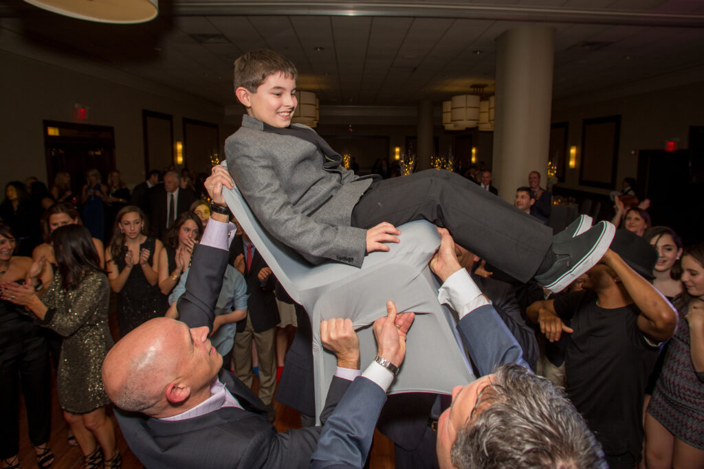 A boy in a gray suit is lifted on a chair by two men during a celebration in a crowded banquet hall.