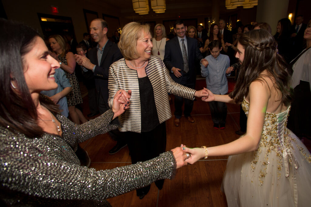Three women hold hands and dance in a circle at an indoor event, with a crowd of people watching and smiling in the background.