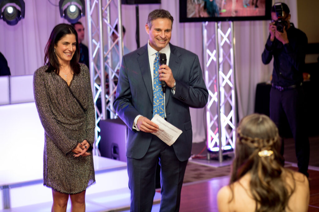 A man in a suit holding a microphone stands beside a woman in a dress, addressing seated guests at an indoor event, with lights and a cameraman in the background.