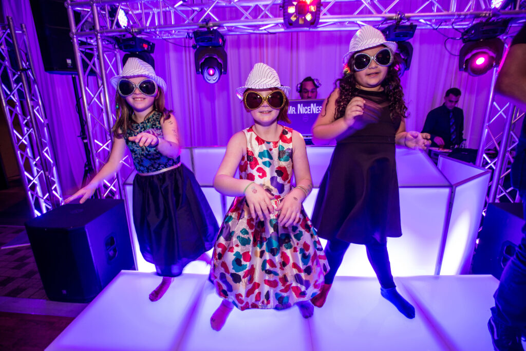 Three young girls wearing white hats and sunglasses dance on a lit stage with purple lighting and a DJ booth in the background.