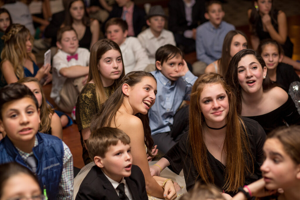 A group of children and teenagers dressed in formal attire sit closely together on the floor, looking forward with engaged and curious expressions.
