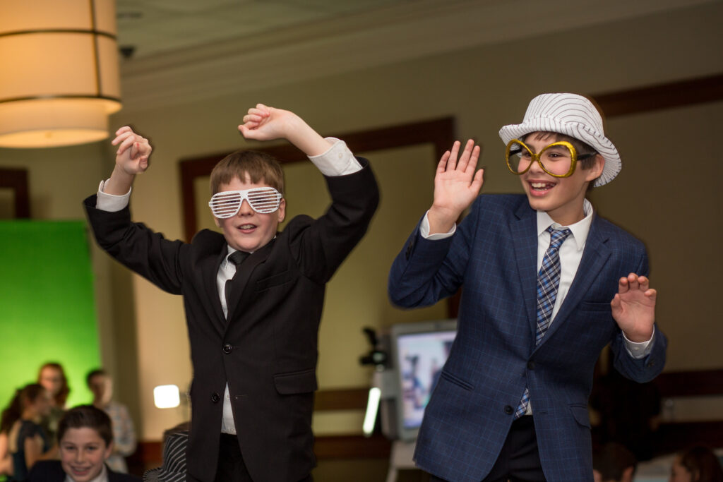 Two boys in suits, wearing playful hats and novelty glasses, dancing indoors at a party or event. Other kids are sitting in the background.