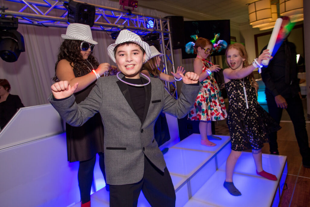 A boy in a gray suit and striped hat dances on a lit platform with several children behind him at a party, all under colorful lighting.