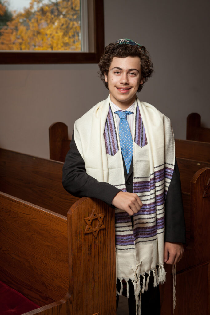 A young person in a suit and tallit stands by a wooden pew with a Star of David, inside a synagogue.