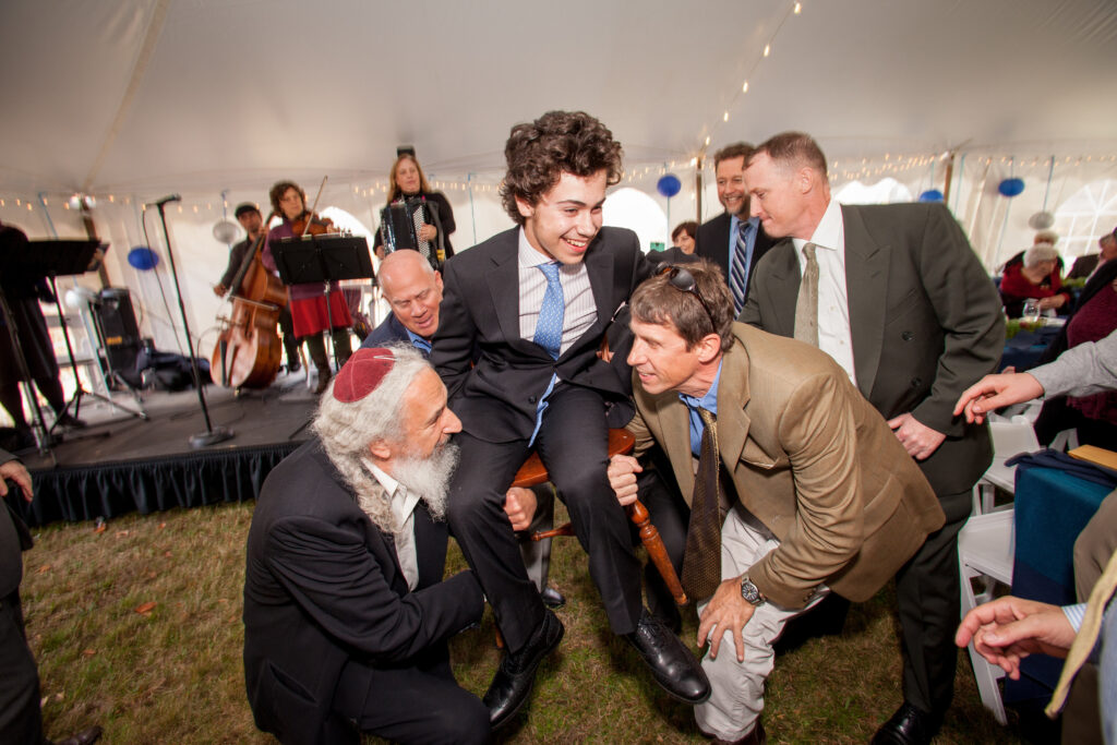 Young man in a suit is lifted on a chair by several men at an indoor celebration with live musicians playing in the background.