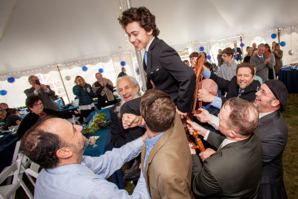 A group of people lift a smiling boy on a chair during a celebration under a tent, while guests watch and applaud in the background.