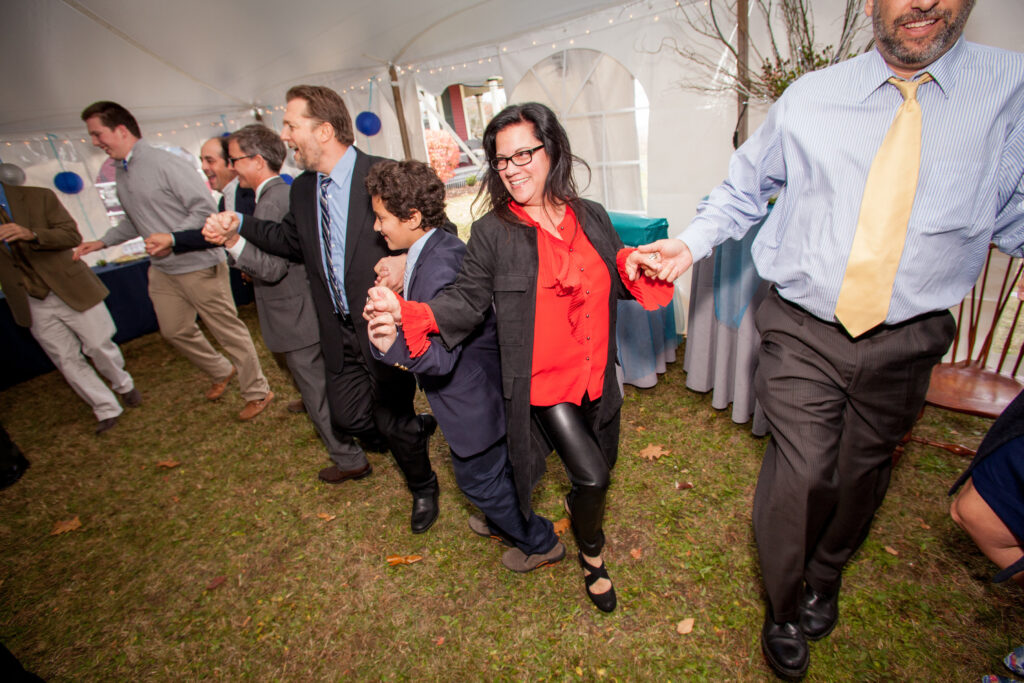 A group of people, dressed in semi-formal attire, hold hands and dance in a line inside a tent on a grassy area during a celebration.