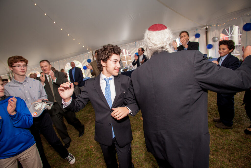 A group of people in formal attire dance and celebrate under a large tent decorated with blue and white balloons.